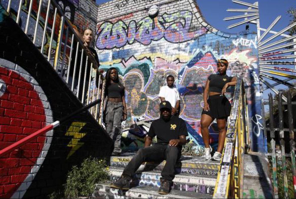Group of people wearing XP London streetwear posing on graffiti-covered wall in an urban setting, showcasing bold street style and creative energy.