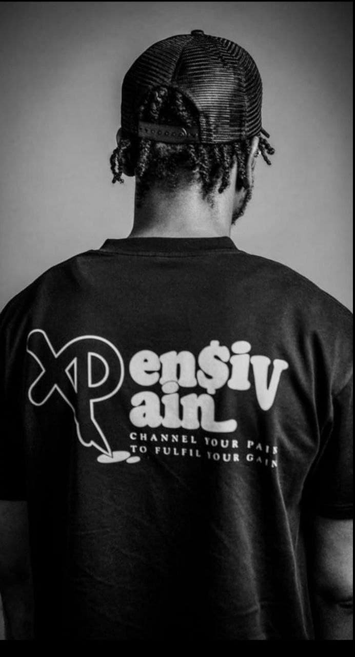 Black and white photography of a model wearing a black XP London ‘XPensiv Pain’ t-shirt with the slogan ‘Channel your pain to fulfil your gain,’ showcasing bold streetwear design.