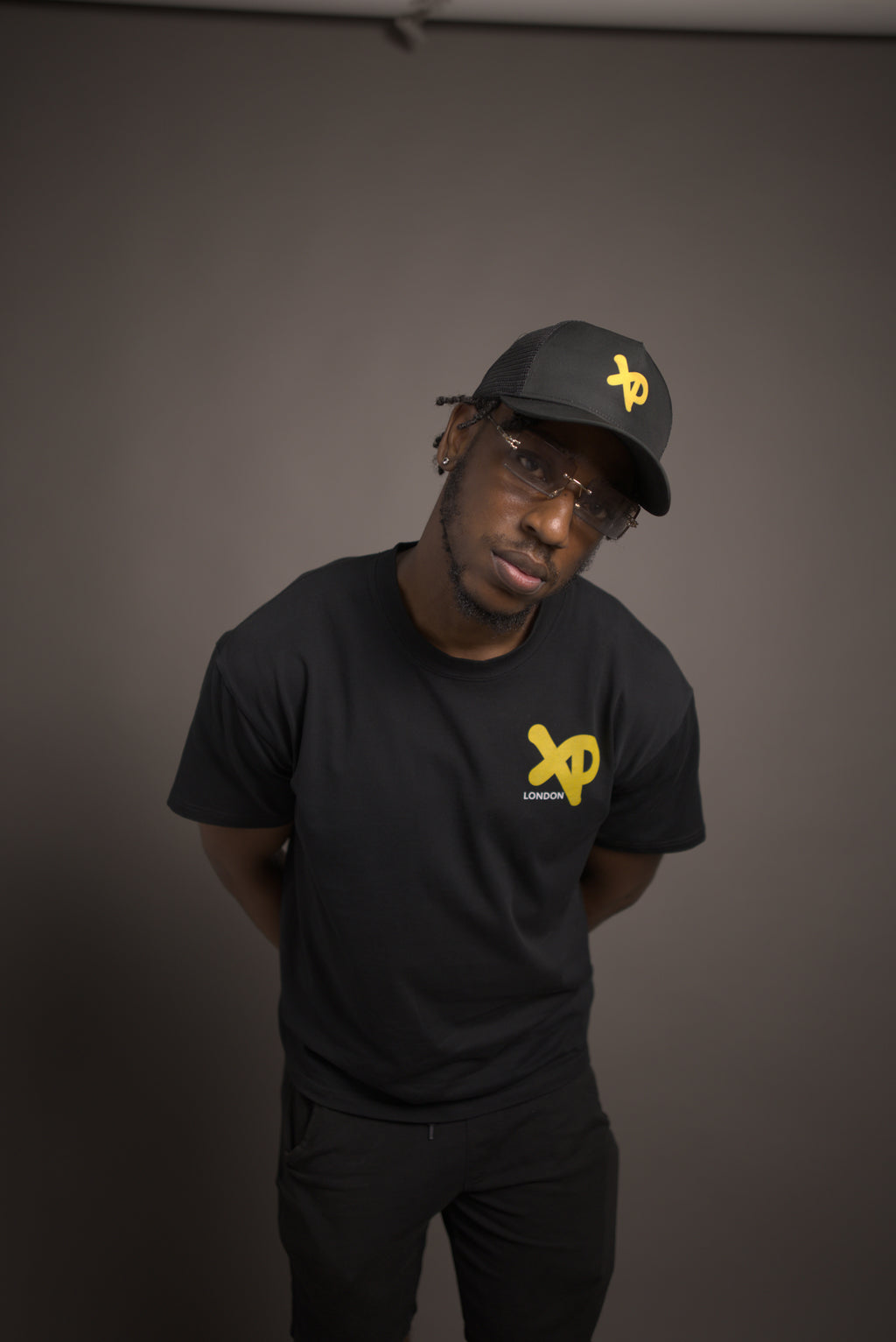 Man wearing black XP London t-shirt and matching cap with yellow logo, posing in minimalist studio streetwear photoshoot.