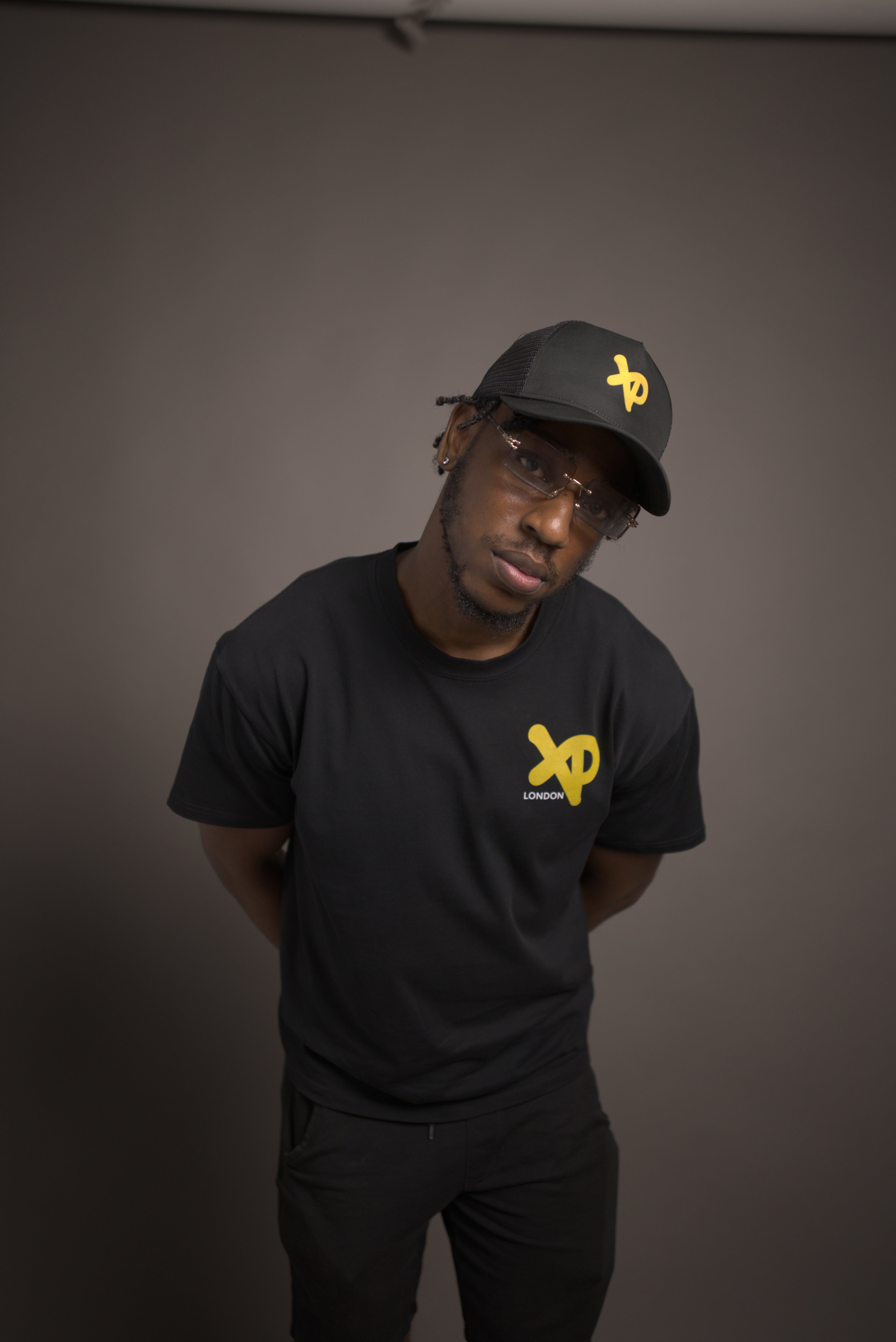 Man wearing black XP London t-shirt and matching cap with yellow logo, posing in minimalist studio streetwear photoshoot.
