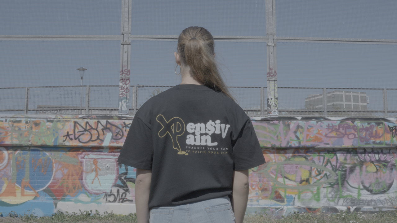 Back view of woman wearing black XP London ‘XPensiv Pain’ t-shirt with yellow and white logo, standing in front of colorful graffiti wall in outdoor urban setting.