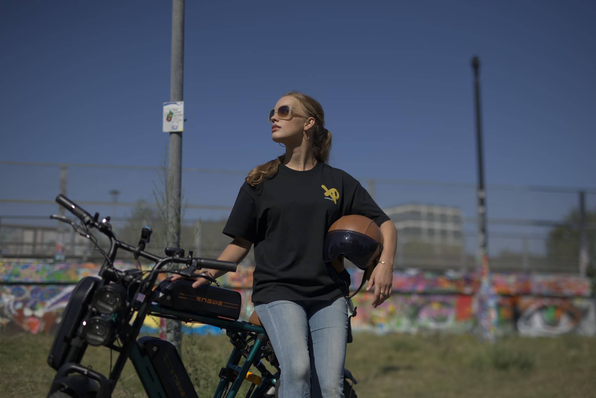 Woman wearing a black XP London t-shirt with yellow logo, holding a helmet beside an electric bike in an urban graffiti park setting.