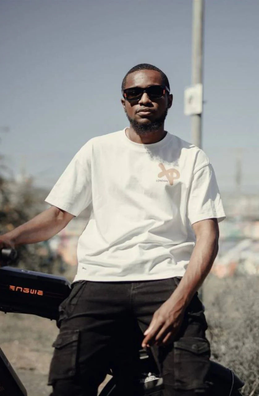 Model wearing a white XP London streetwear t-shirt and black cargo pants sitting on a bike in an urban outdoor setting, showcasing casual modern fashion.