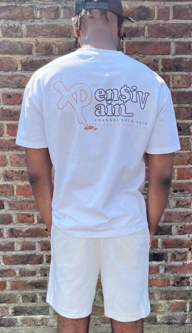 Man wearing a white XP London ‘XPensiv Pain’ t-shirt and shorts, standing against a brick wall, showcasing minimalist summer streetwear style.