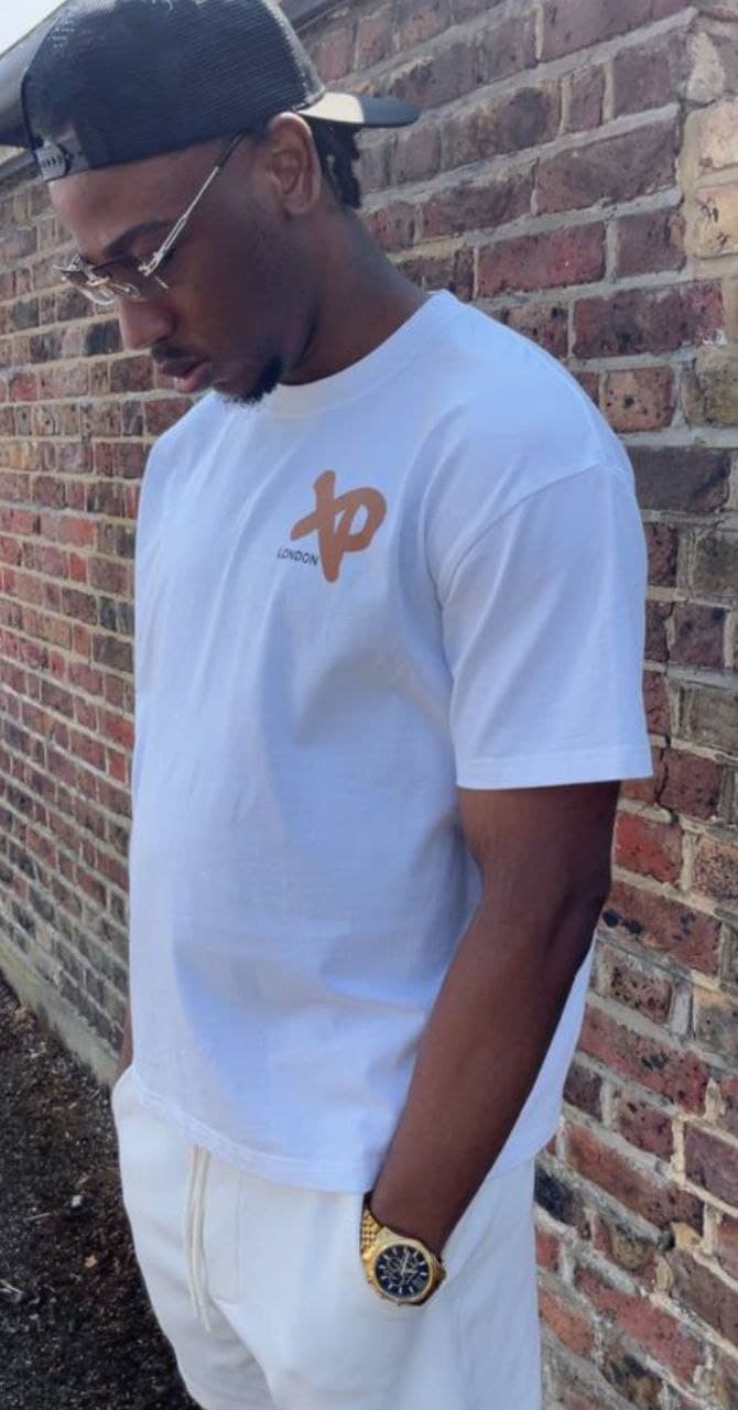 Man wearing a white XP London t-shirt and black cap standing against a brick wall, showcasing clean minimalist streetwear style.