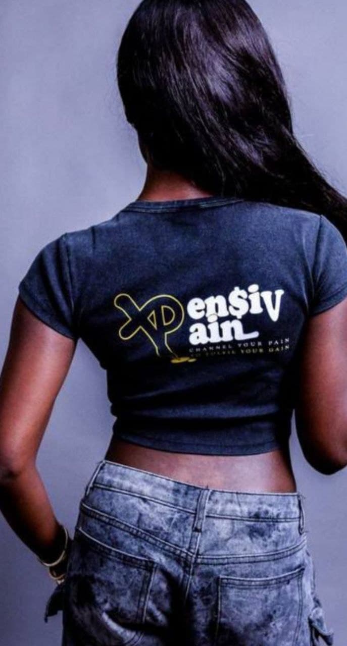 Back view of woman wearing a charcoal-black XP London ‘XPensiv Pain’ cropped t-shirt with yellow and white logo, styled with grey cargo jeans in studio streetwear photoshoot.