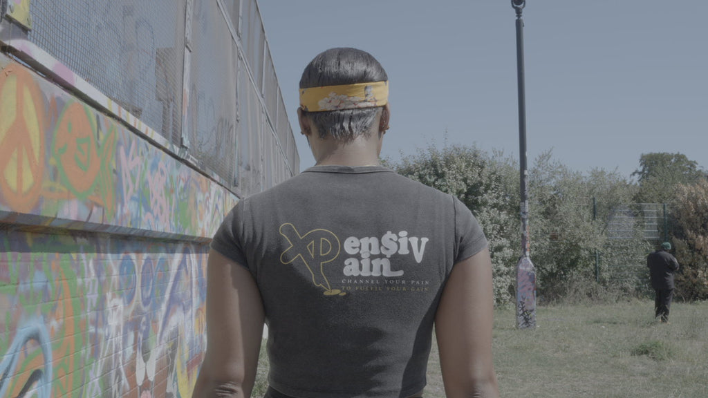 Back view of woman wearing black XP London ‘XPensiv Pain’ crop top with yellow and white logo, standing beside graffiti-covered wall in outdoor urban setting.