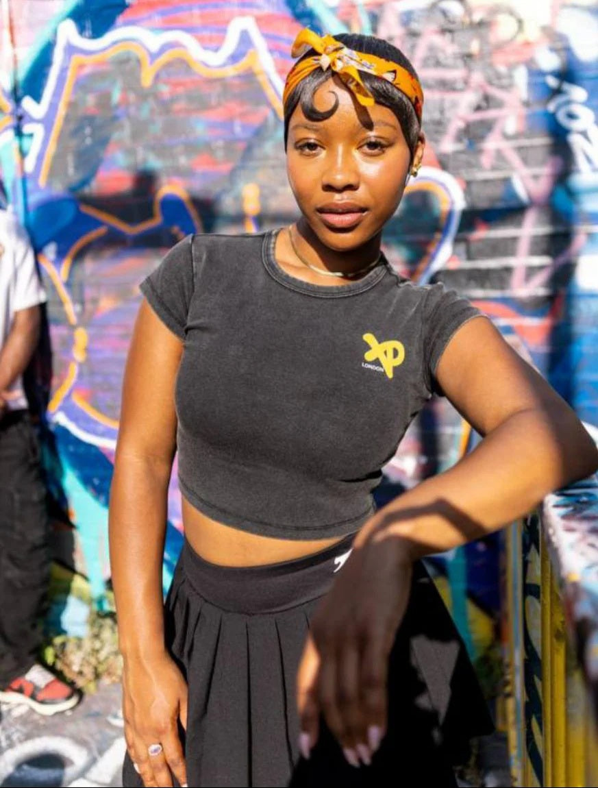 Woman wearing a charcoal-black XP London crop top and pleated skirt posing confidently against a colorful graffiti wall, representing bold urban streetwear style.