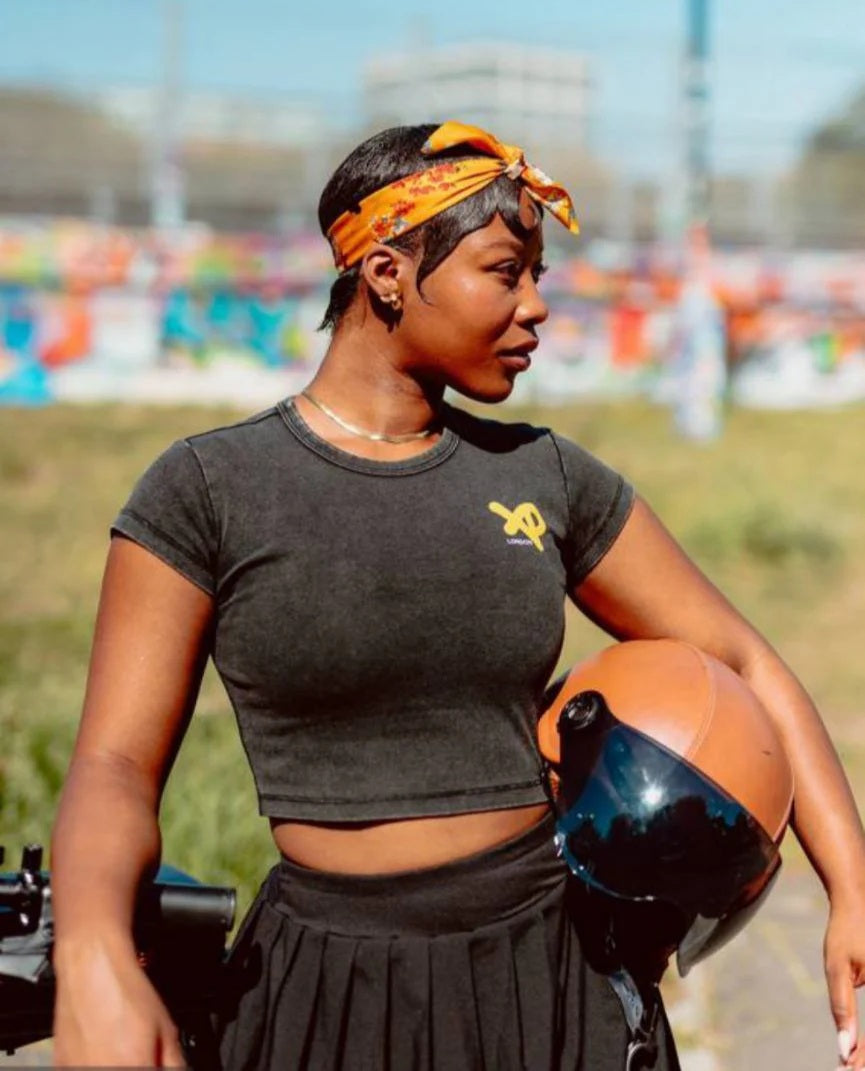 Woman wearing a black XP London crop top and pleated skirt with an orange headband, holding a motorcycle helmet in an urban graffiti-filled setting.