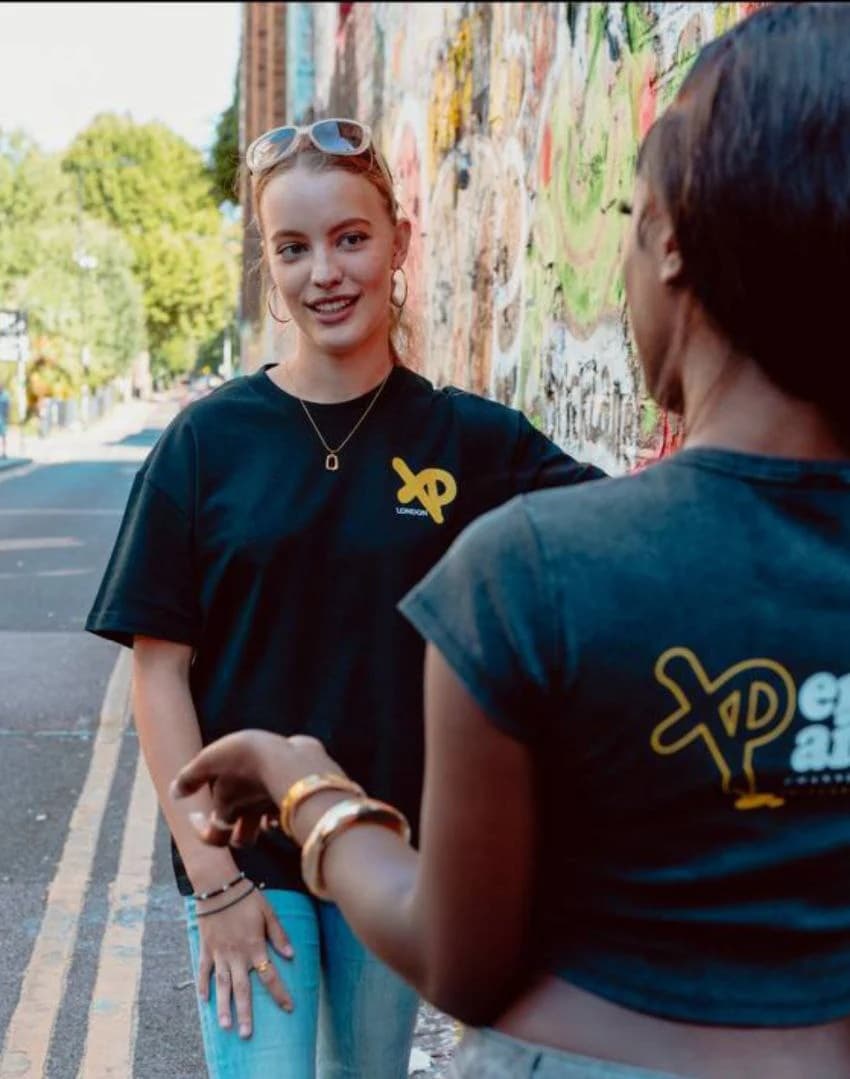 Two women wearing XP London streetwear t-shirts talking by a graffiti-covered wall on a city street, showcasing urban fashion and community vibes.