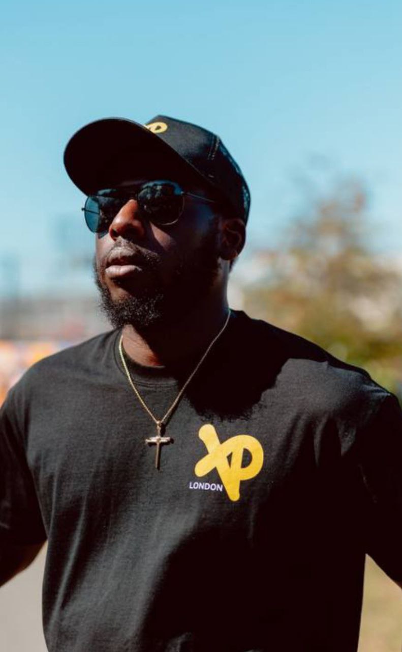 Male model wearing XP London black t-shirtr and cap under clear blue sky in urban settng