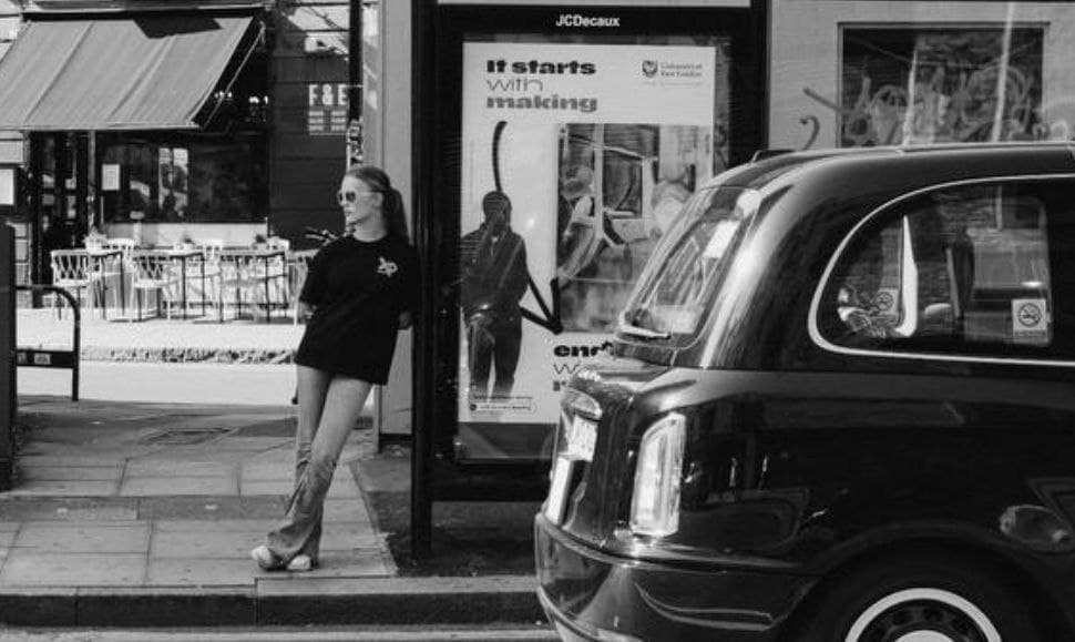 Model wearing XP London streetwear leaning at a city bus stop in London beside a classic black cab, captured in black and white for an urban fashion vibe.