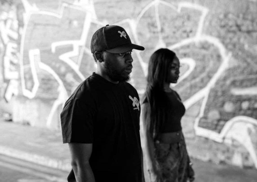 Two models wearing XP London streetwear standing in front of a graffiti-covered wall, black and white urban fashion photography highlighting minimalist street style.