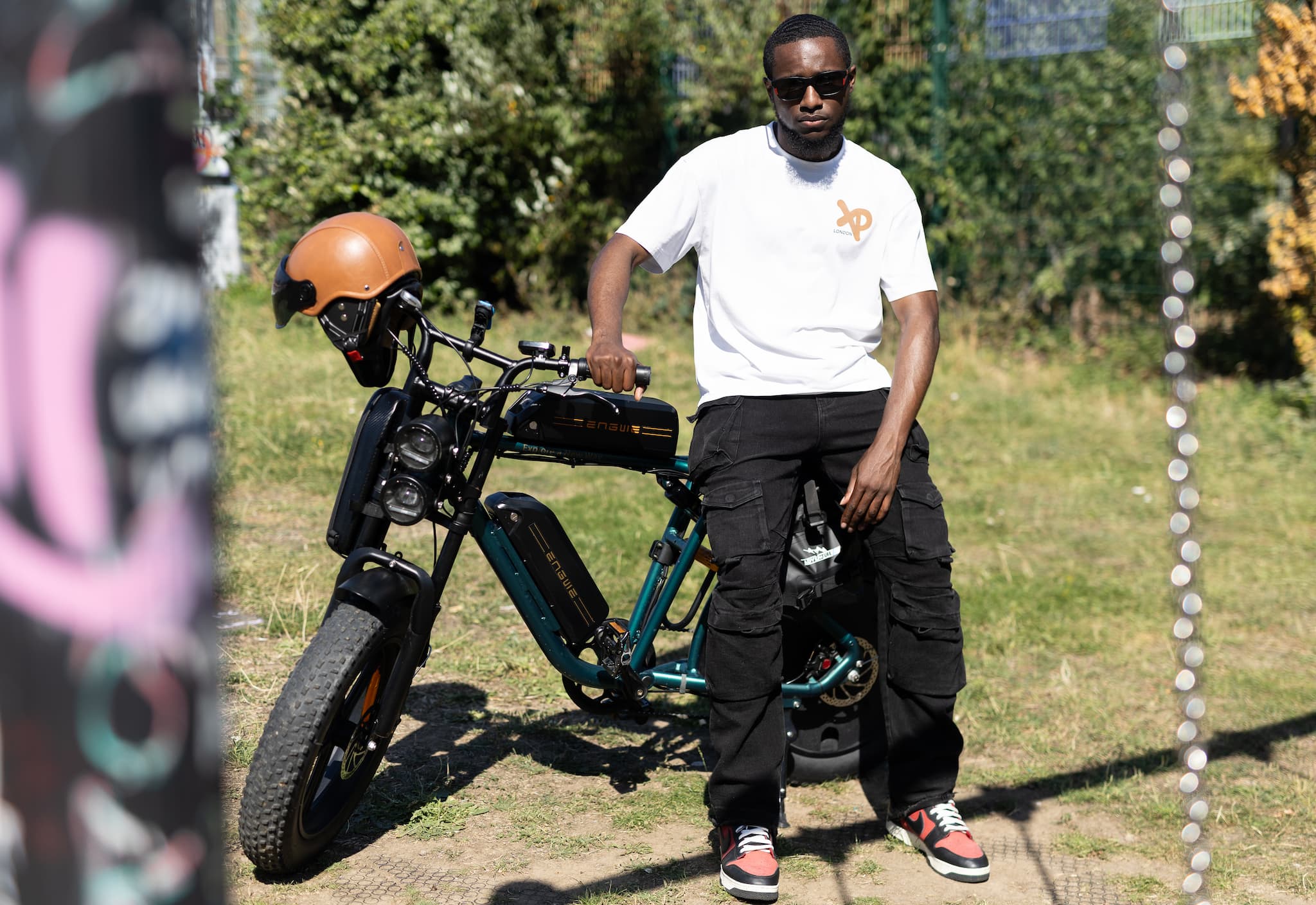 Model in white XP London t-shirt sitting on black electric bike outdoors in bright sunlight