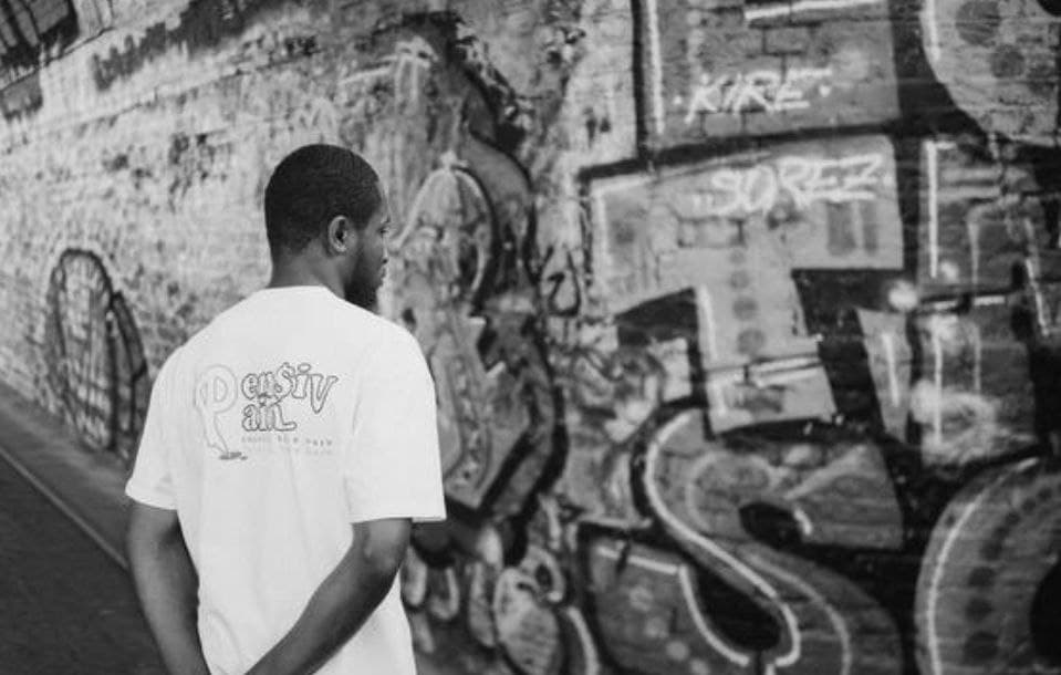 Model wearing a white XP London ‘XPensiv Pain’ t-shirt walking along a graffiti-covered brick wall in black and white, showcasing minimalist streetwear style.